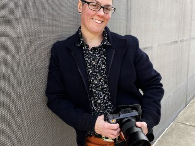 Photo of Jess T. Dugan with a black blazer, dark patterned shirt and orange patterns holding a camera along a grey wall