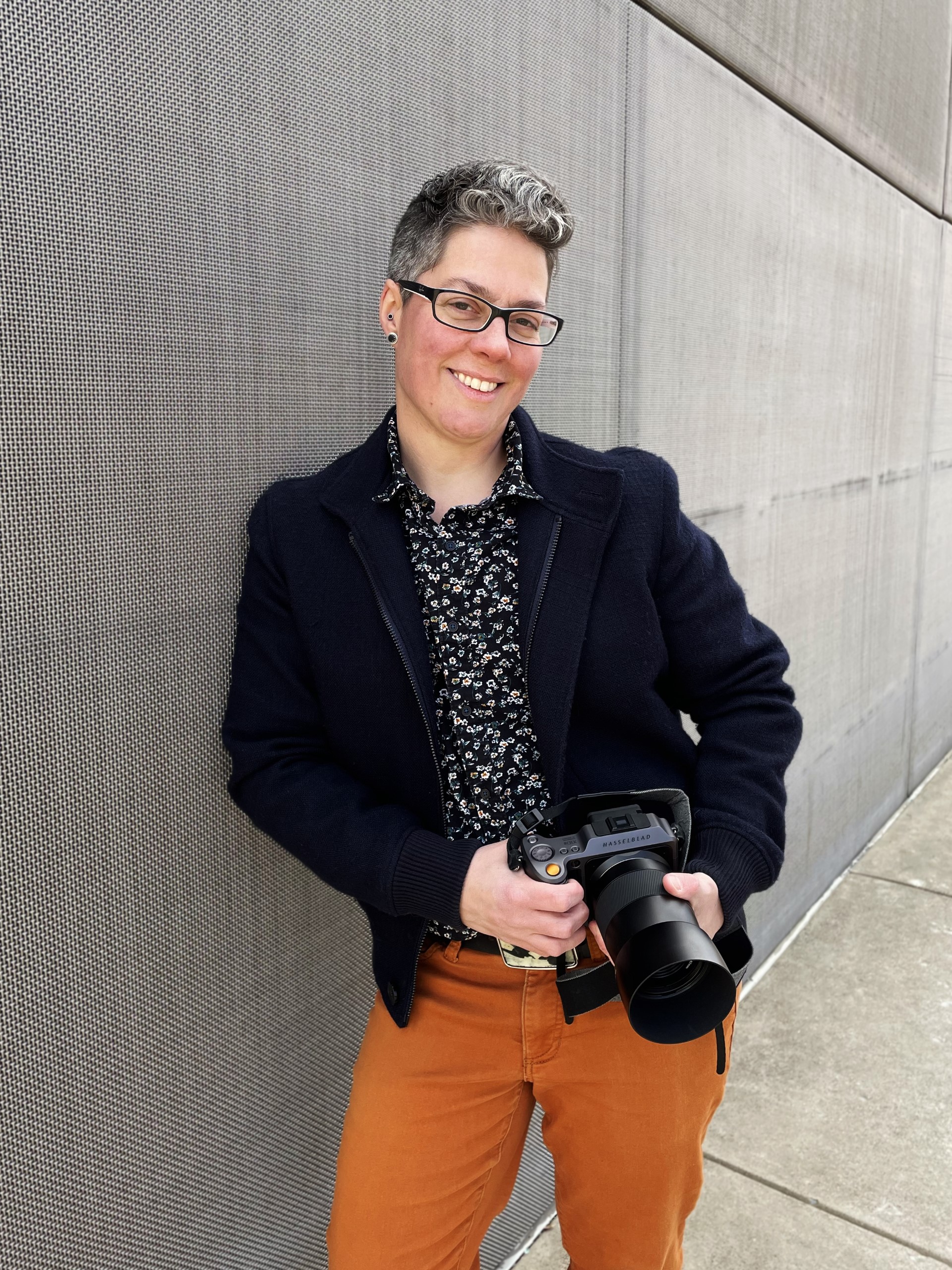 Photo of Jess T. Dugan with a black blazer, dark patterned shirt and orange patterns holding a camera along a grey wall