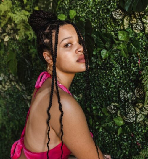 Portrait of Kierah KIKI King along a plant wall. KIKI is wearing a magenta sleeveless top.